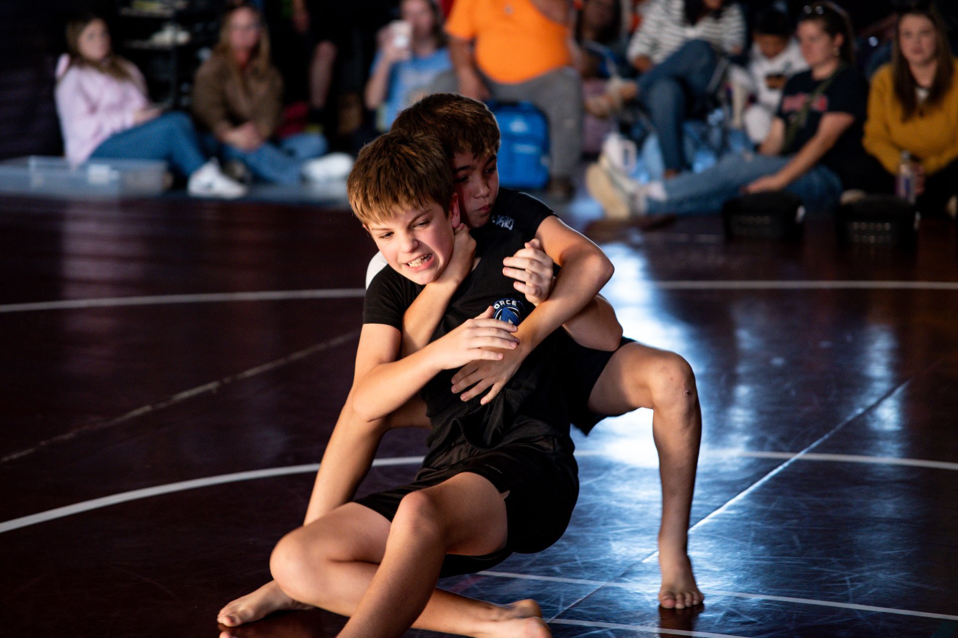 Two wrestlers in black grappling on a mat, audience in background.