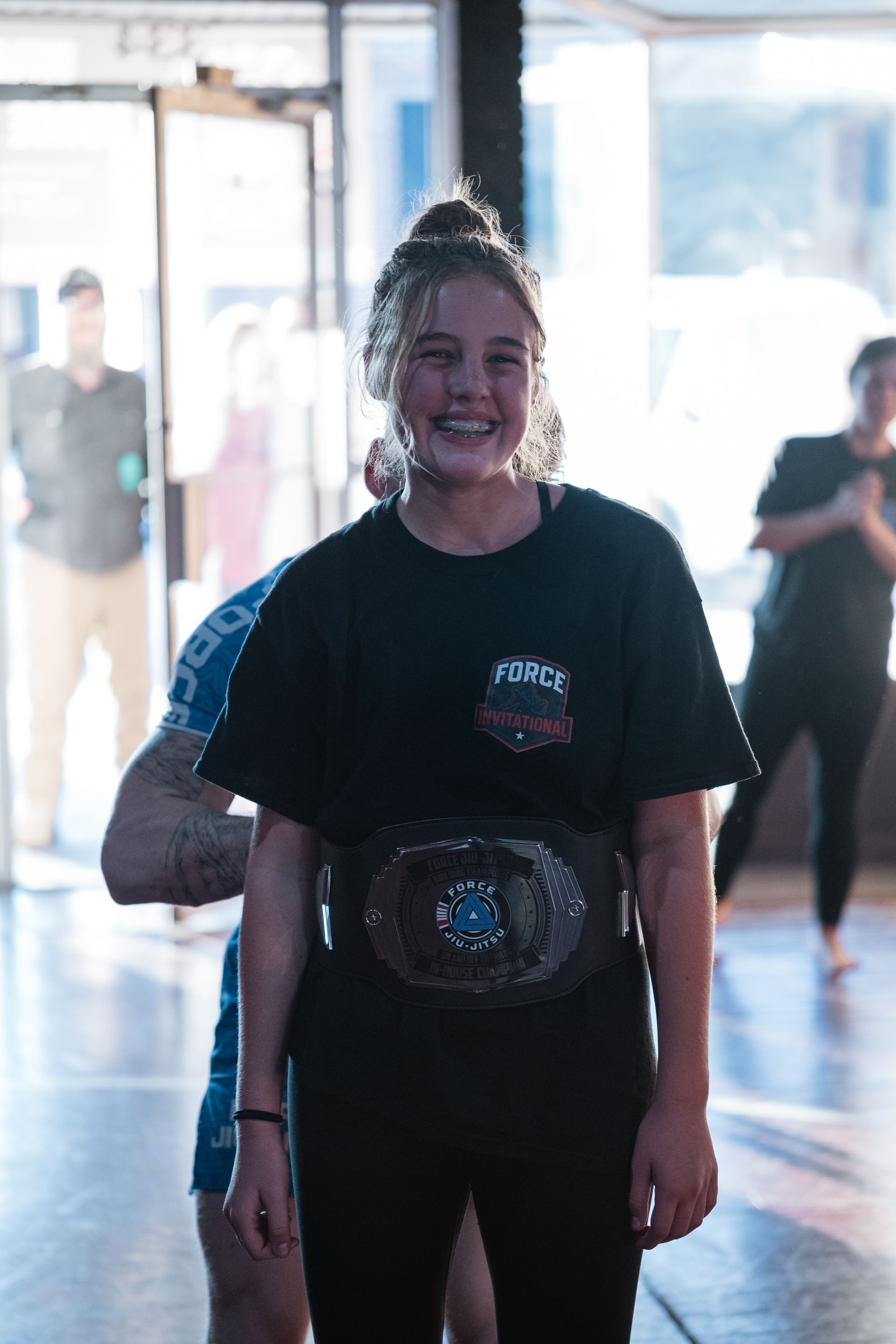Girl in black grappling gear facing away from another person, in a gym that's wrapping a championship belt around her after she won her jiu-jitsu match. Other people are in the background.