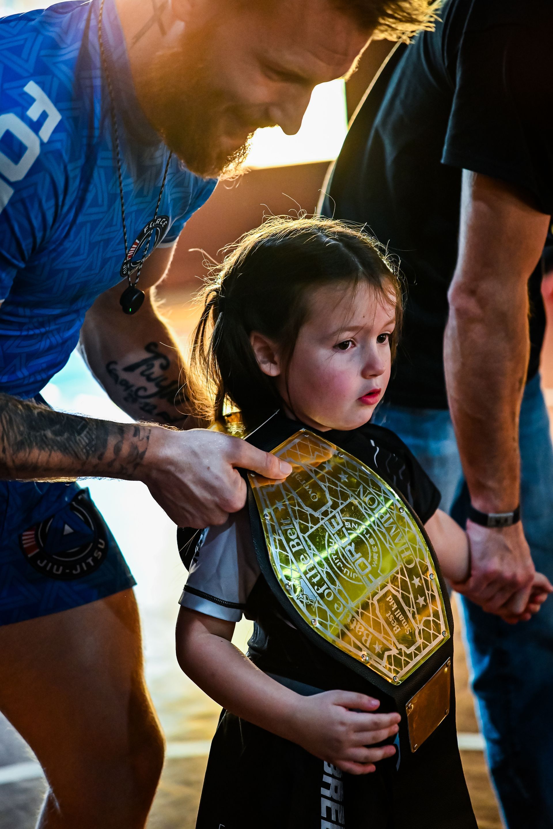 Man adjusts a championship belt on a small child. They stand outside, backlit by the sun.
