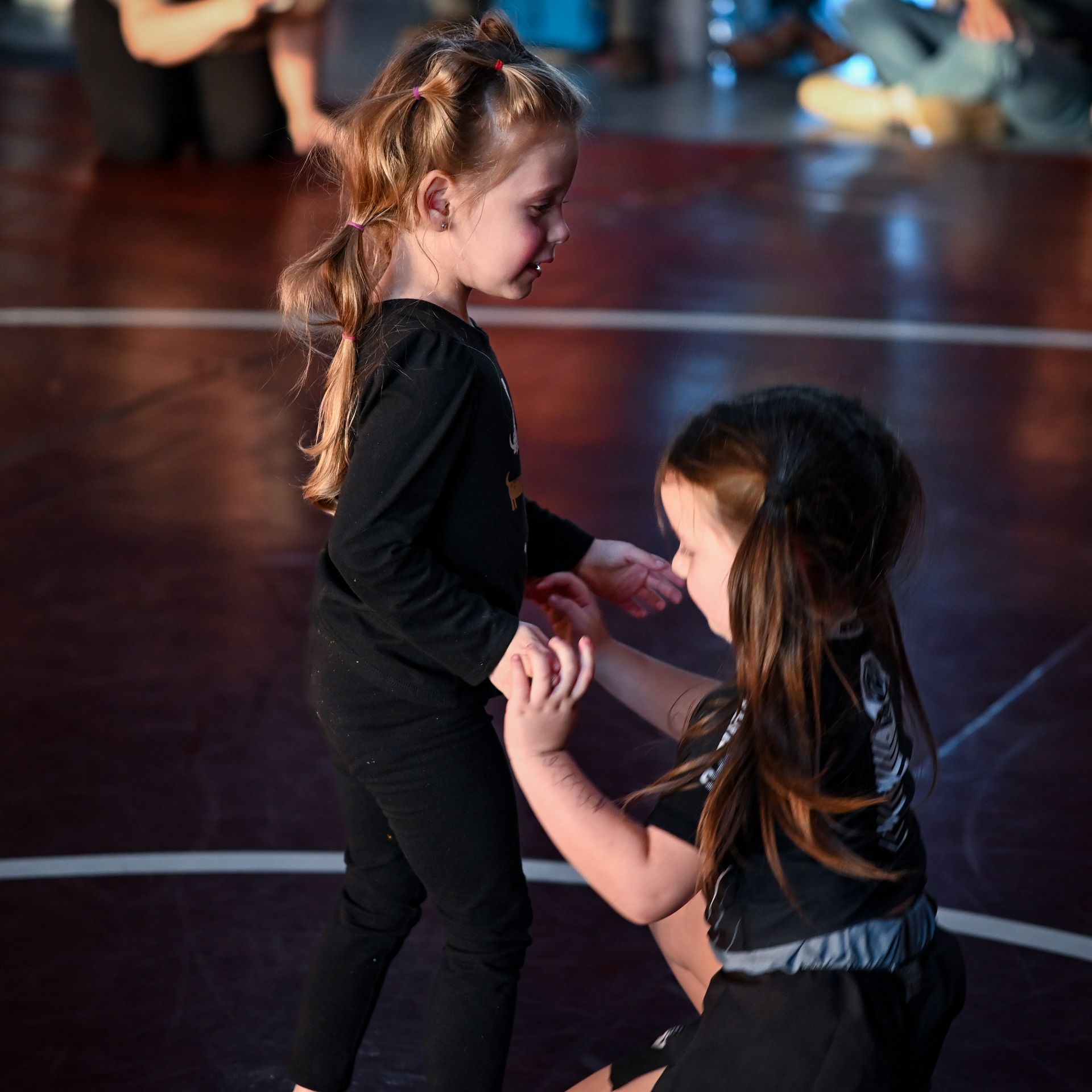 Two young children in black outfits, one kneeling, dancing on a gym floor.
