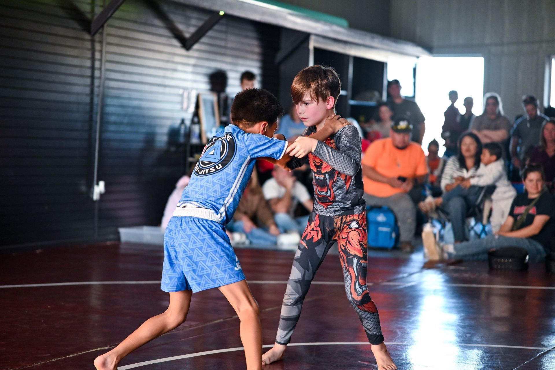 Two young wrestlers grappling on a red mat during a match. Spectators watch in the background.