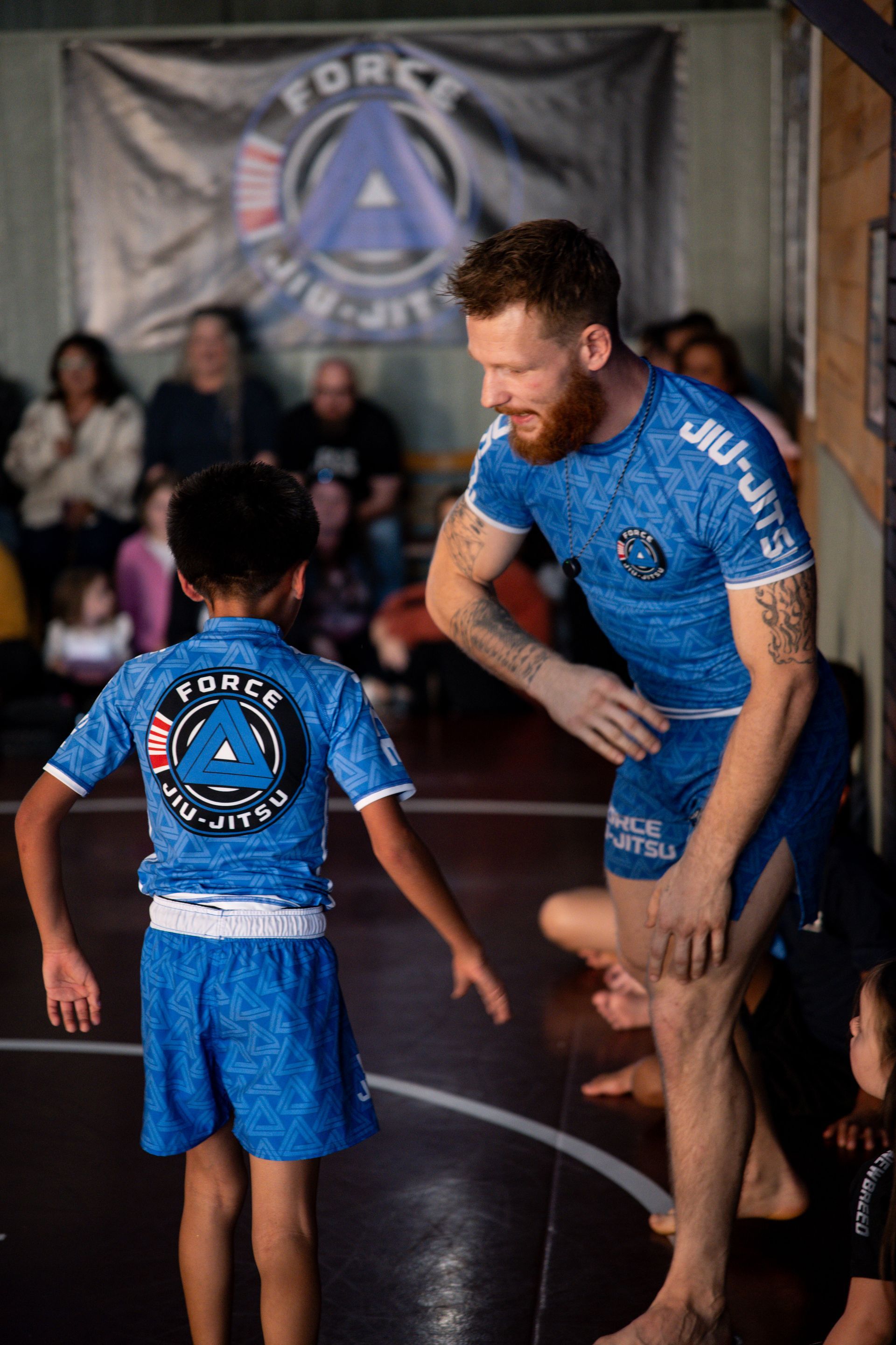 Adult in blue Jiu-Jitsu gear congratulates a child in matching gear; both stand on a mat, indoors.