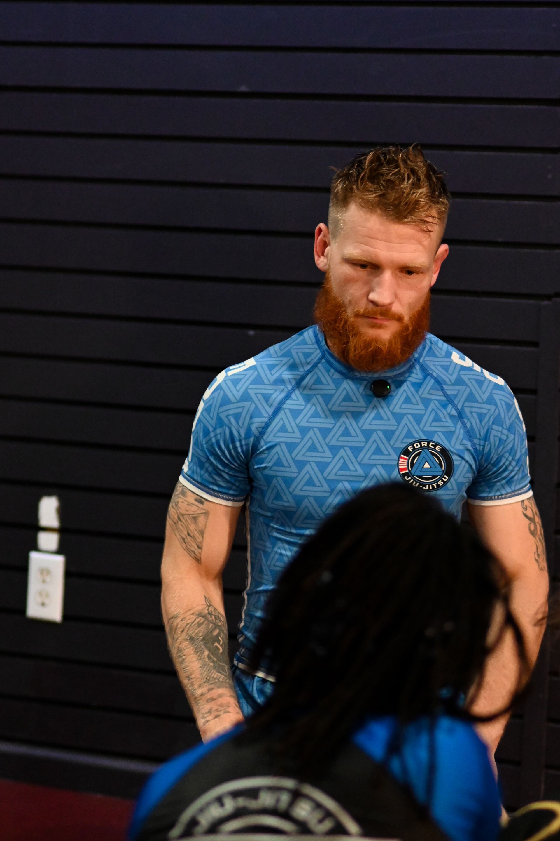 Man with red beard, blue shirt, standing, looking down, gym setting.