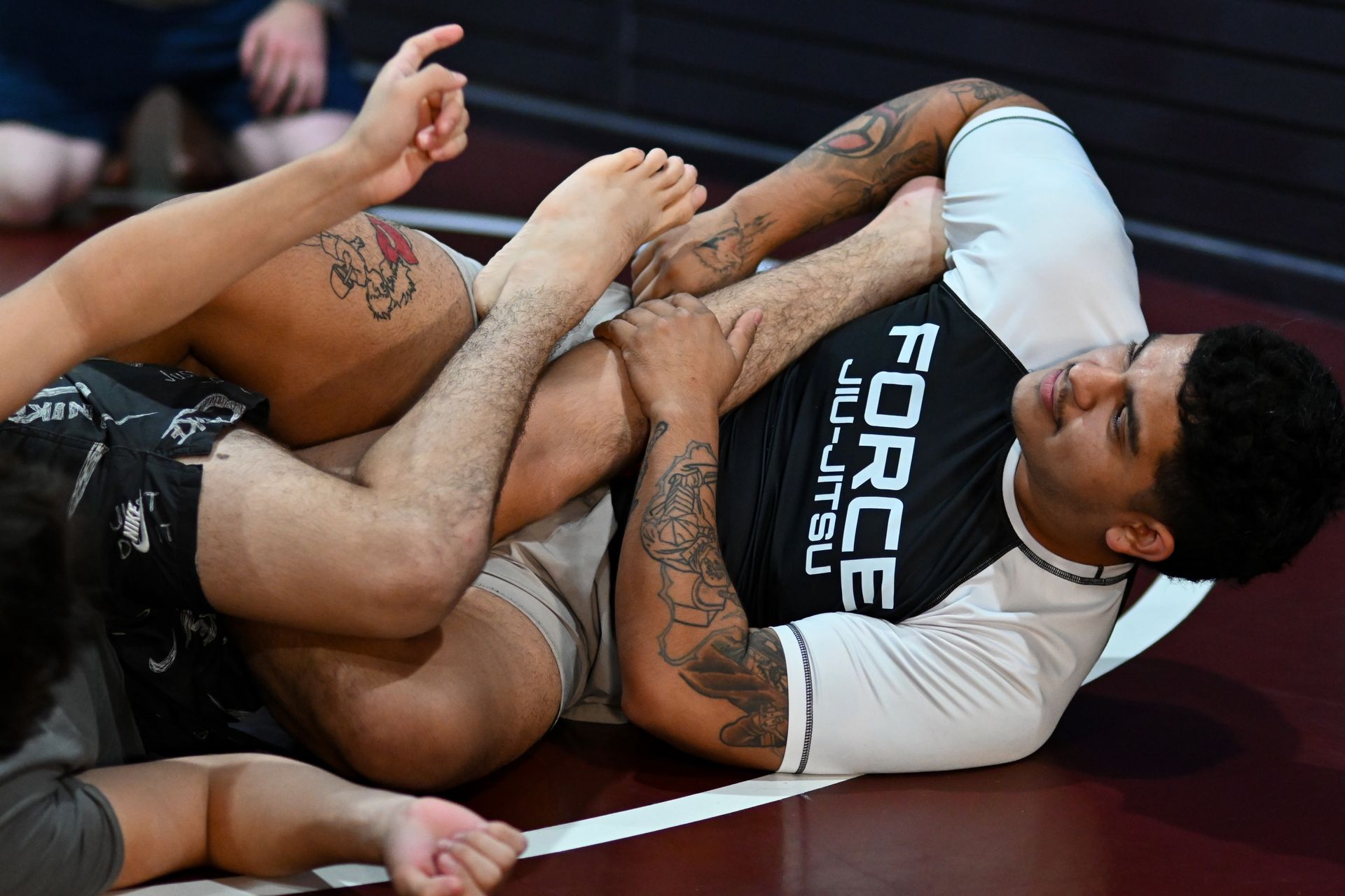 Two people grappling on a mat, one in a Force Jiu-Jitsu shirt, locked in a leg lock.