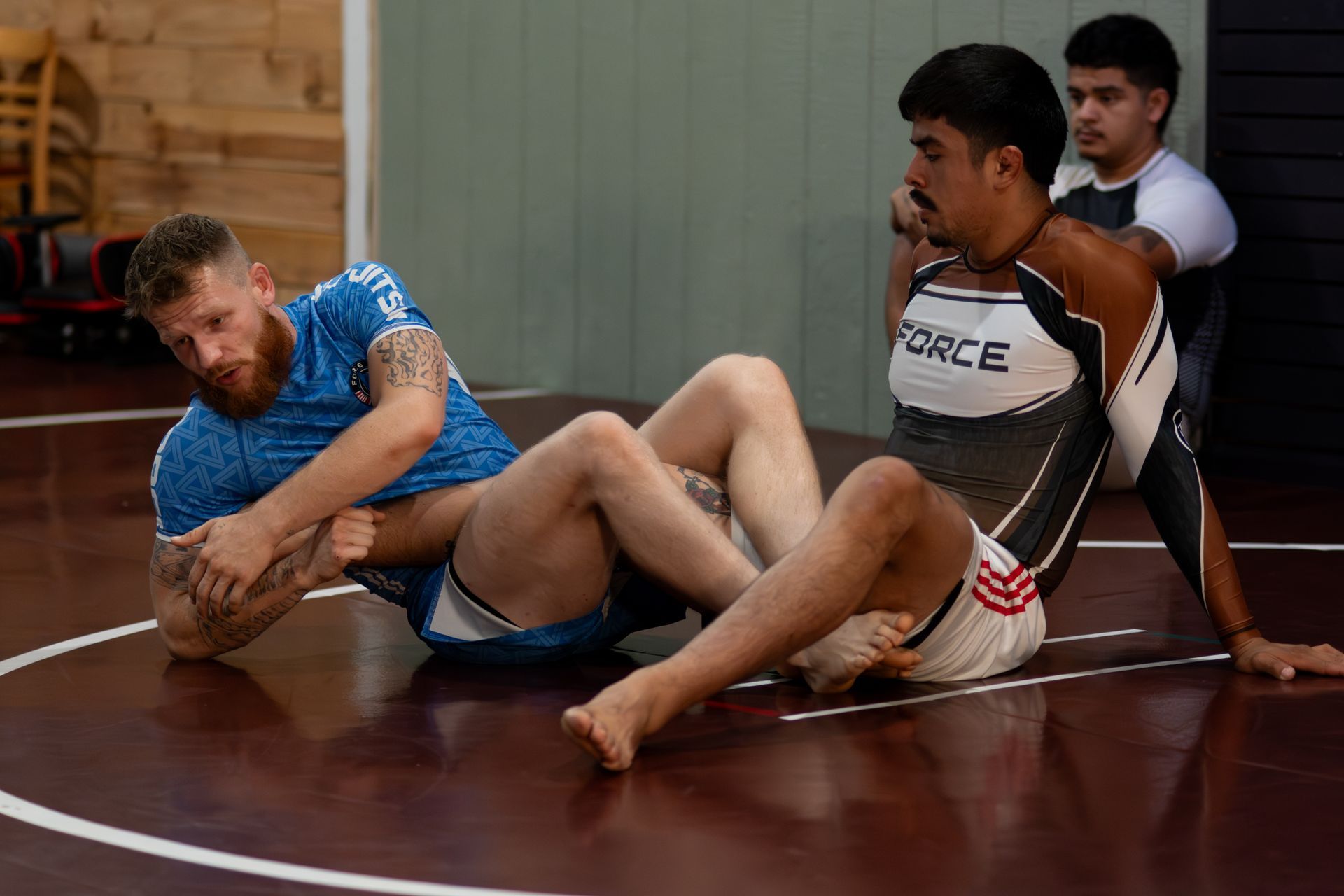 Grappling: Two men in athletic gear on a mat, one in a hold, a third person observing.