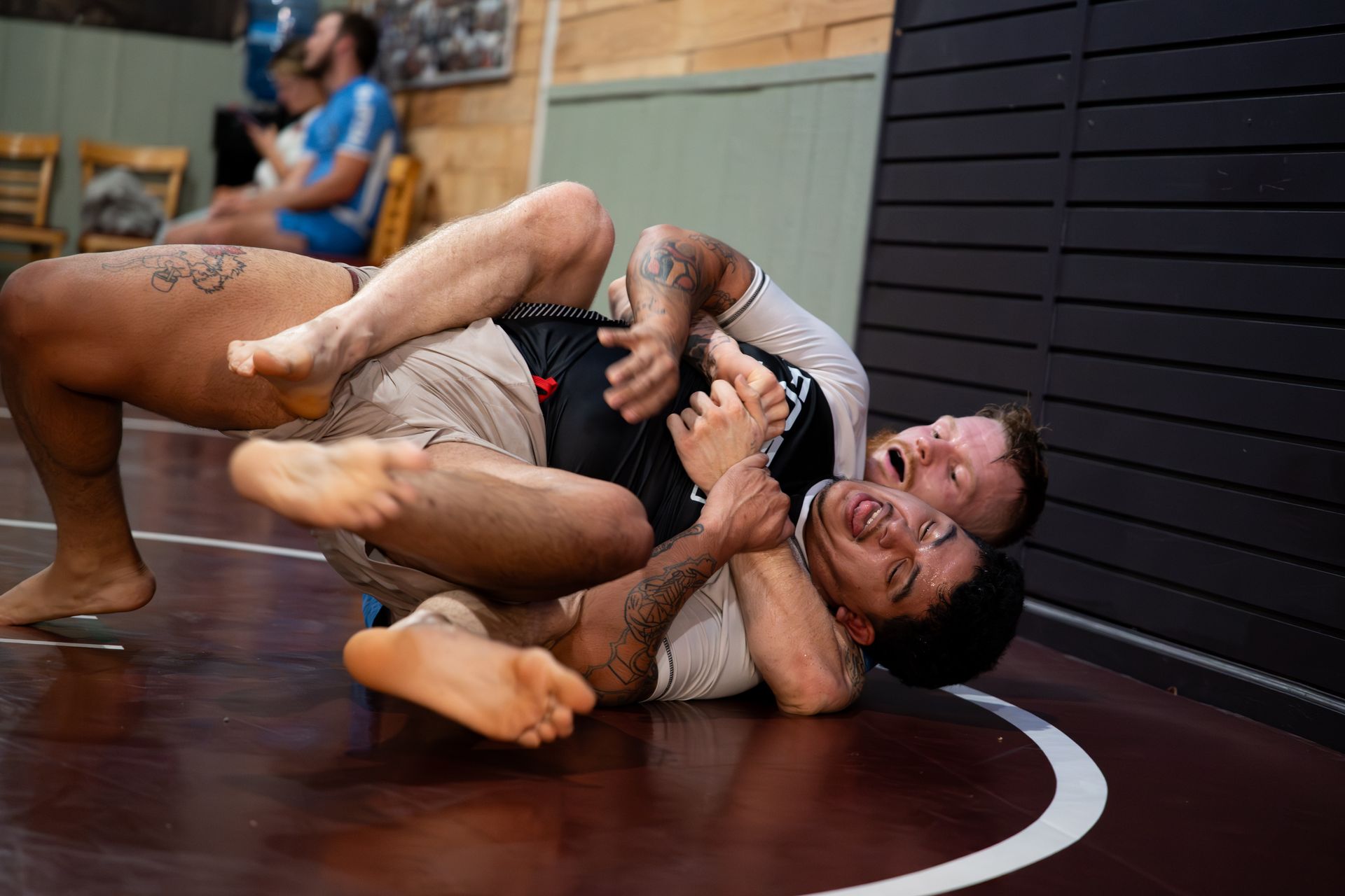 Two men grappling on a mat; one is on top in a headlock, the other struggling with an open-mouthed expression.