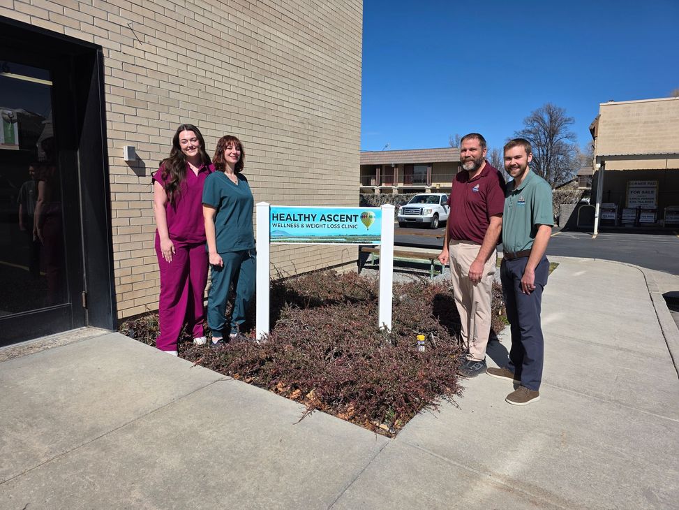 Four people stand smiling by a sign for “The Wellness Center” outside a tan brick building on a sunny day.