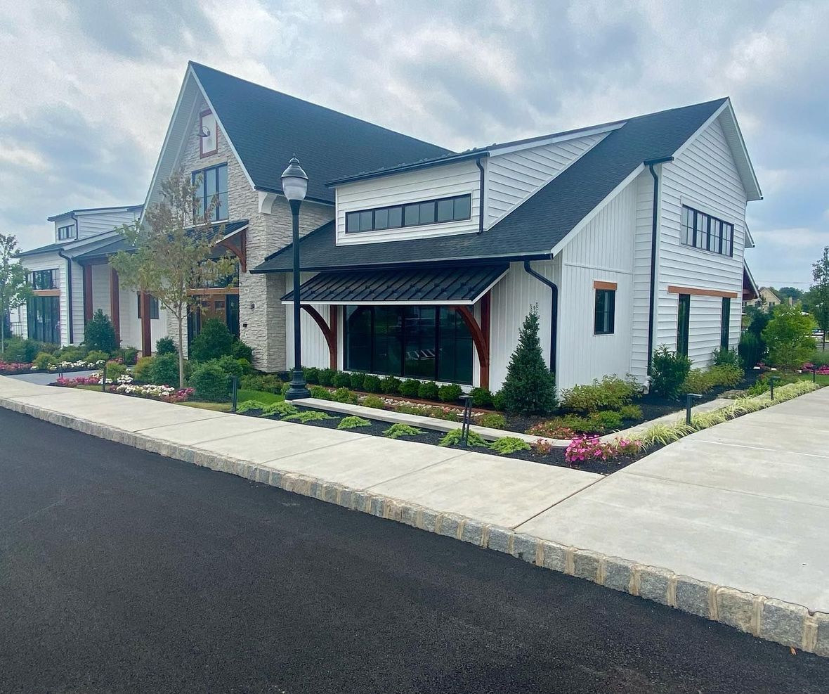 White farmhouse-style building with black roof, stone accents, and manicured landscaping, next to a paved road.