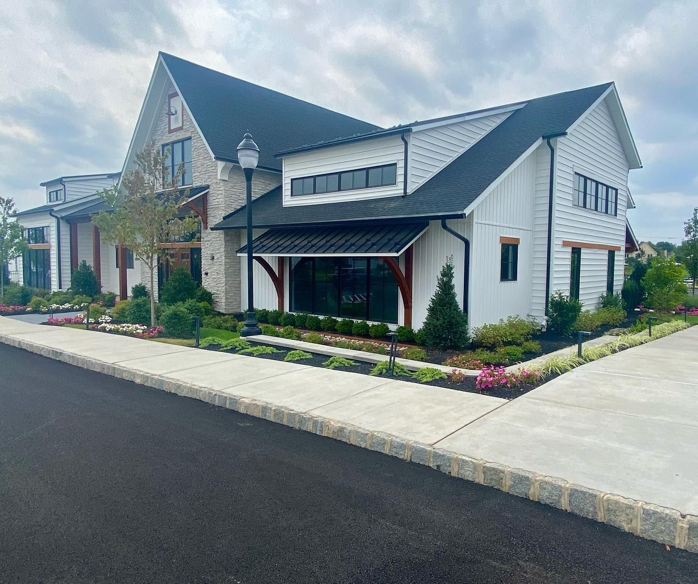 White farmhouse-style building with black roof, stone accents, and manicured landscaping, next to a paved road.