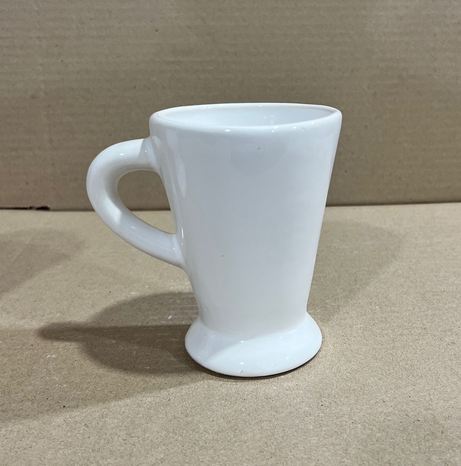 Taza de café de cerámica blanca con asa redondeada, sobre superficie neutra.
