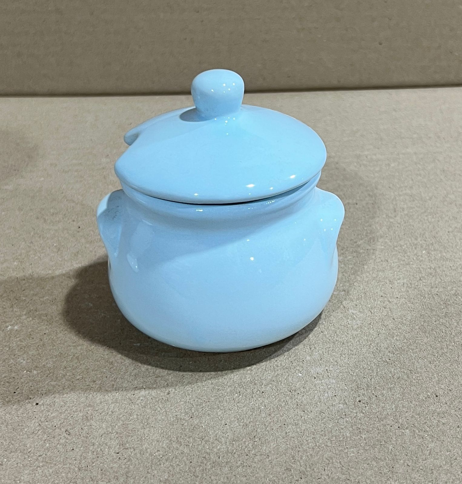 Azucarero de cerámica de color azul claro con tapa y pequeñas asas, colocado sobre una superficie de color marrón claro.