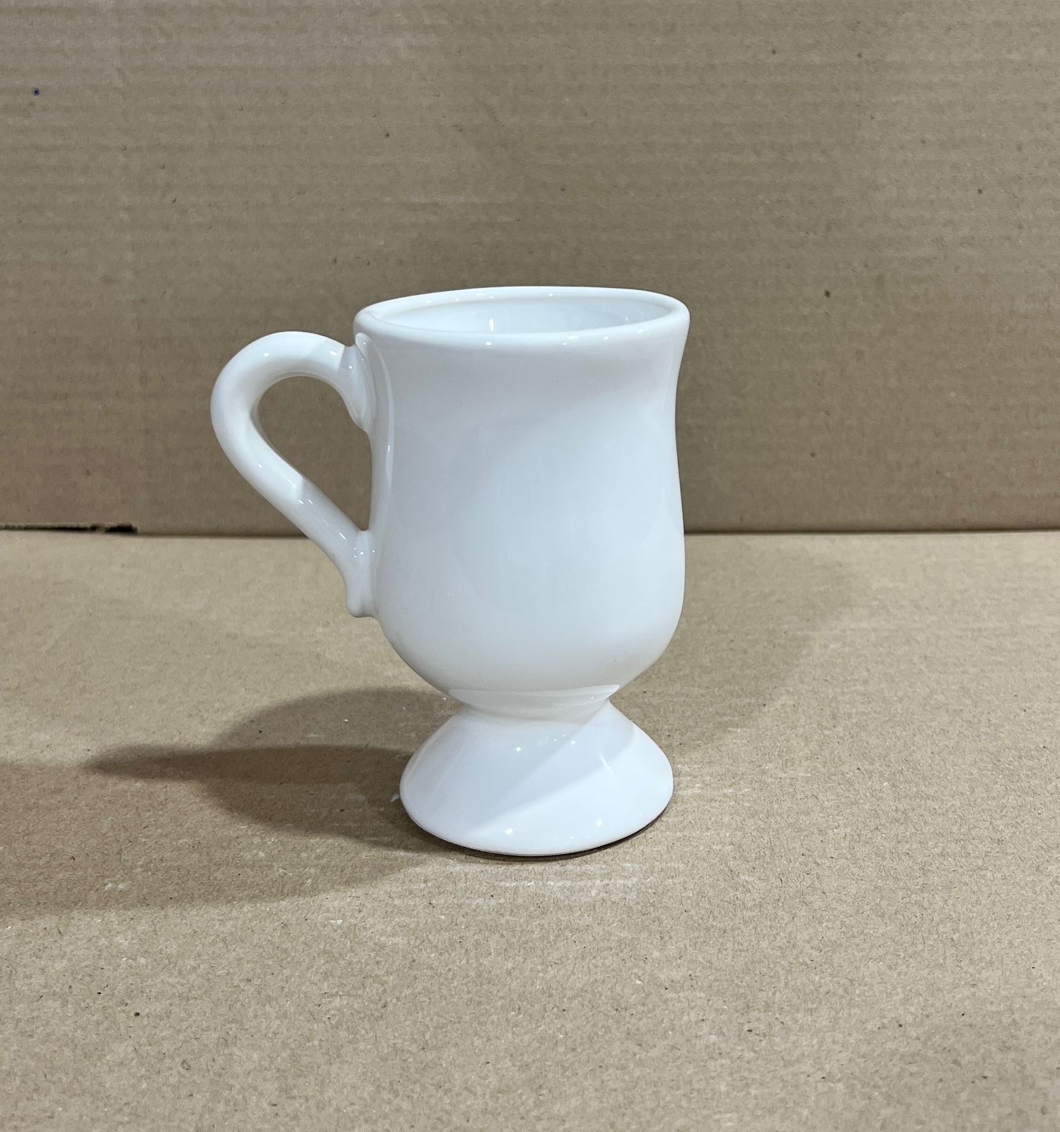 Taza de café con pedestal de cerámica blanca y asa curva, colocada sobre una superficie de color marrón claro.