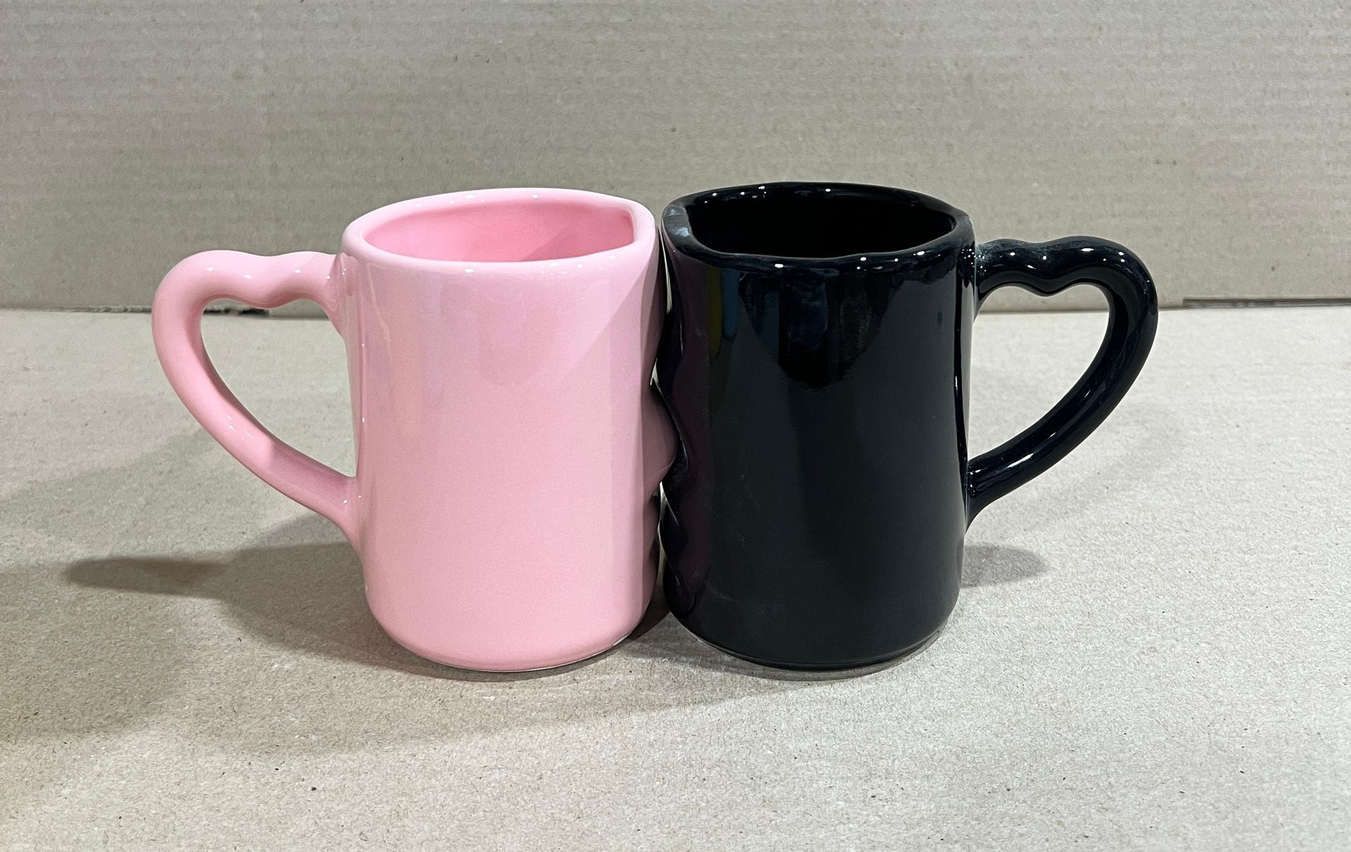 Dos tazas de cerámica: una rosa y otra negra, conmovedoras, con asas en forma de corazón, sobre un fondo liso.