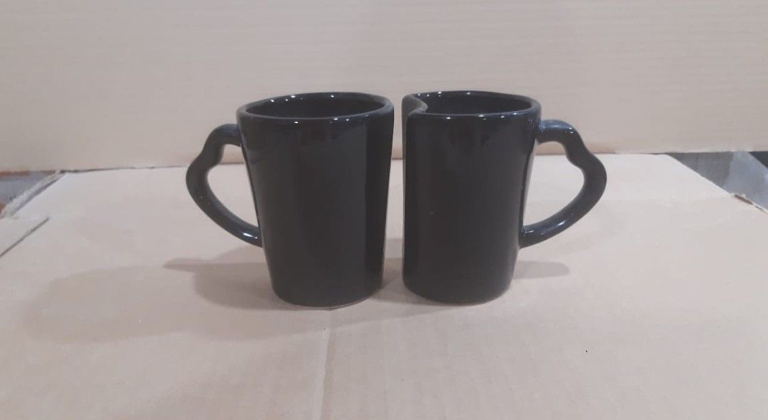 Dos tazas altas de cerámica negra, una al lado de la otra, cada una con asas en forma de corazón, sobre una superficie beige.