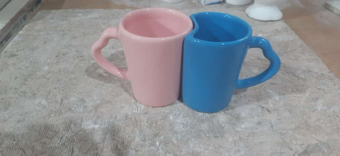 Dos tazas altas de cerámica negra, una al lado de la otra, cada una con asas en forma de corazón, sobre una superficie beige.