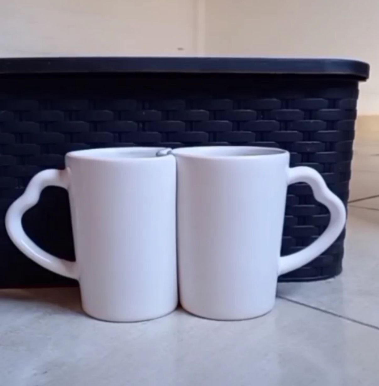 Dos tazas blancas con asas en forma de corazón se encuentran una al lado de la otra frente a una canasta tejida de color negro.