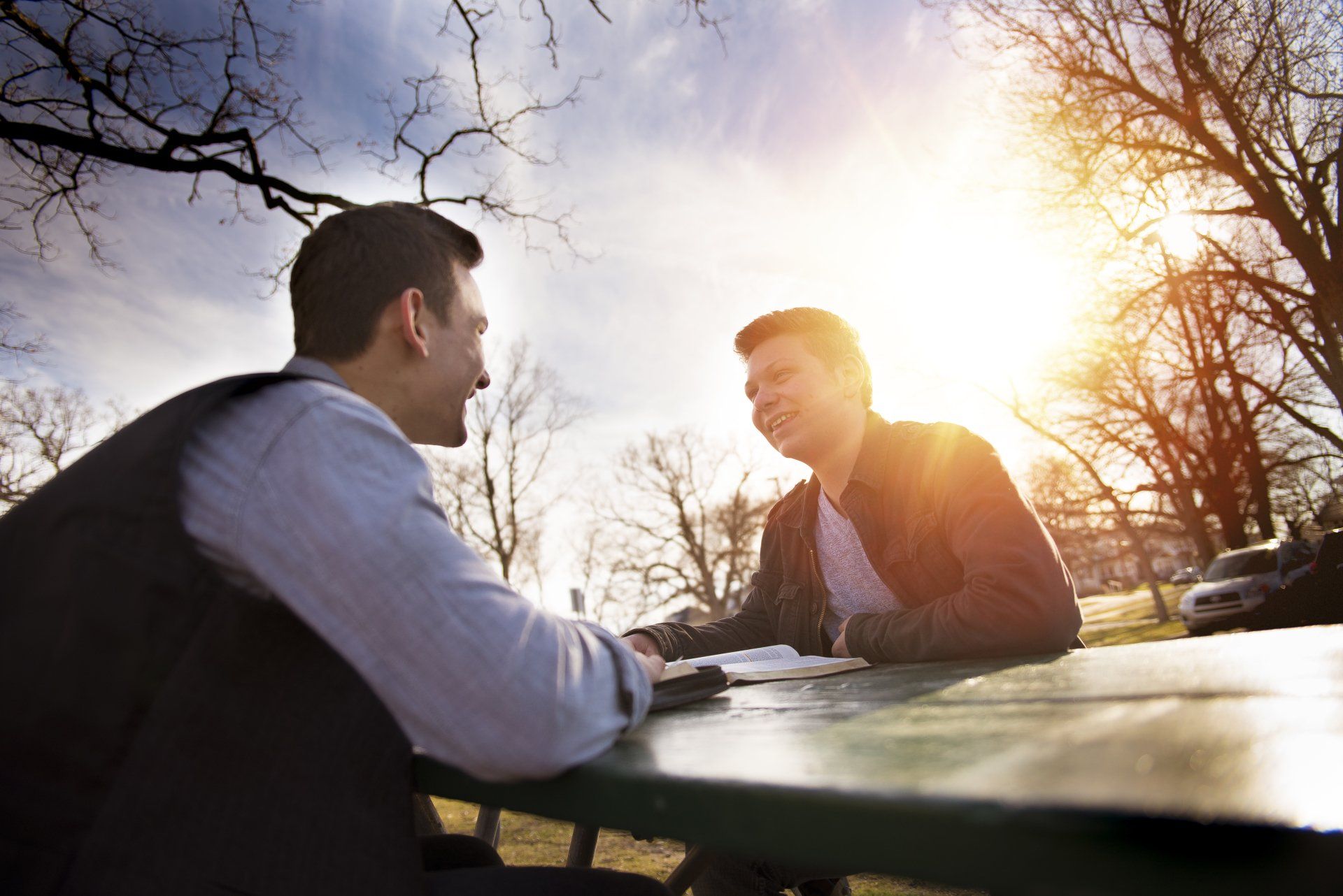 Two people have a conversation at an outdoor table while sunlight glows in the background.