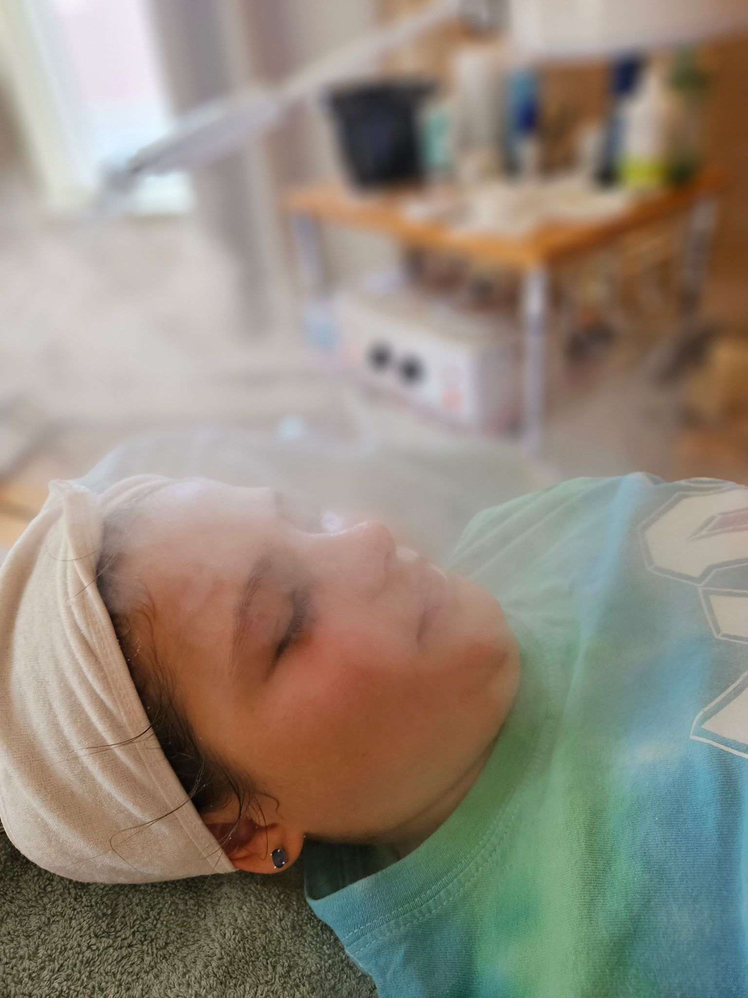Person with a towel on their head receiving facial steam, eyes closed.