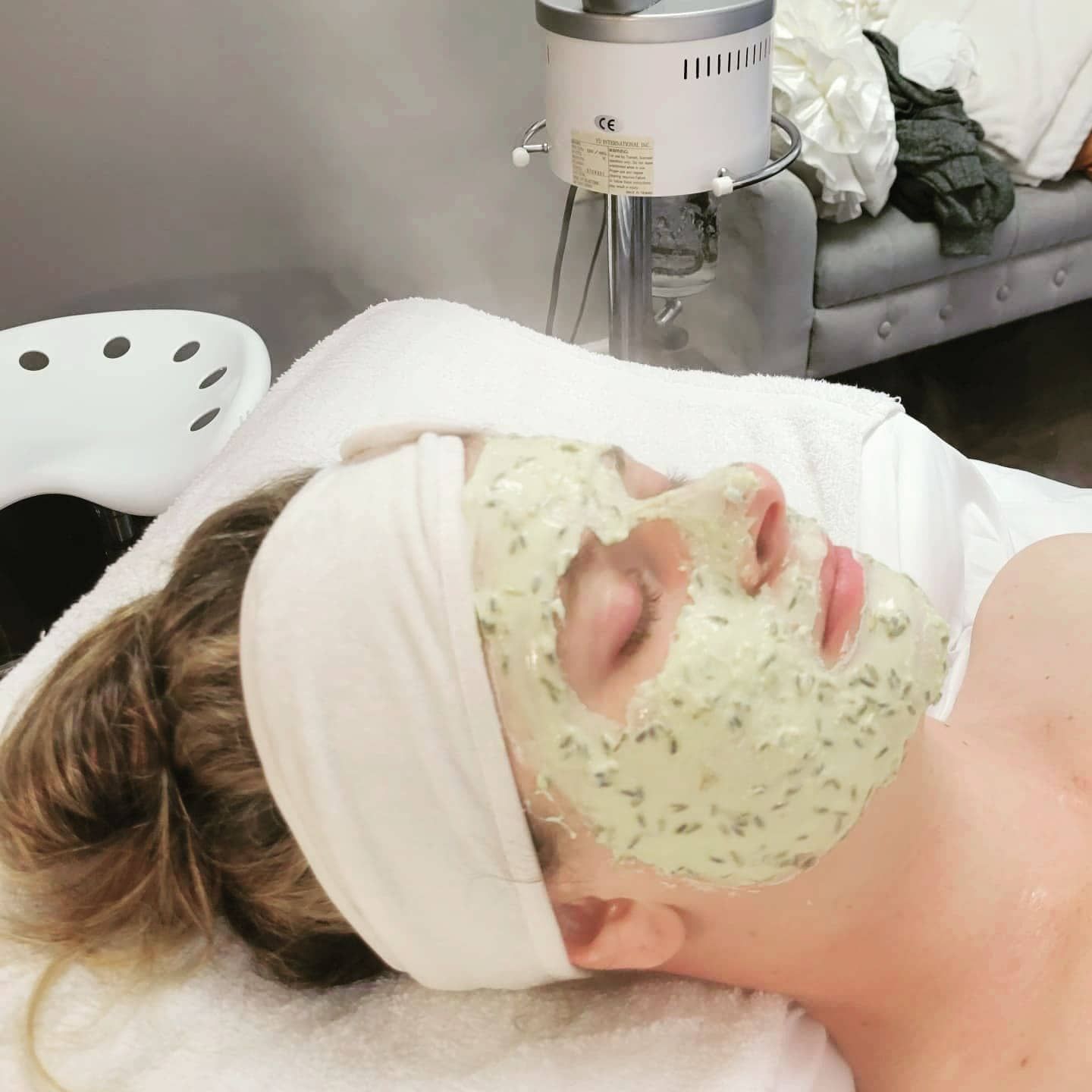 Woman with a face mask lying down, eyes closed, wearing a white headband in a spa setting.
