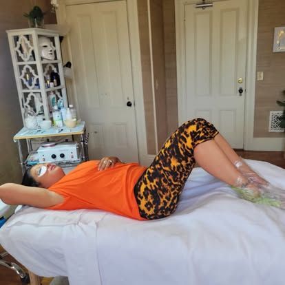 Woman lying on a massage table, wearing an orange shirt and patterned leggings, with eyes covered, in a treatment room.