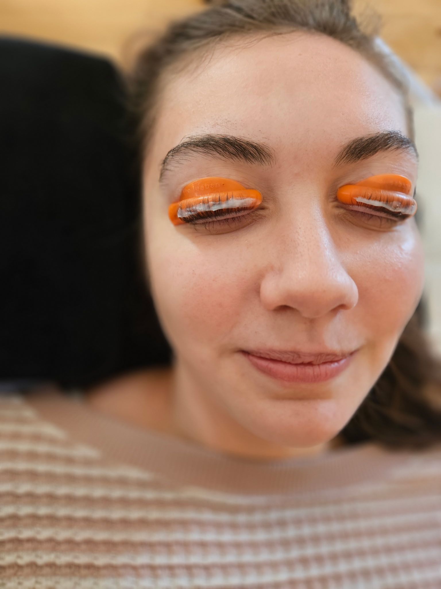 Woman with orange lash lift shields on her eyelids, eyes closed, smiling.