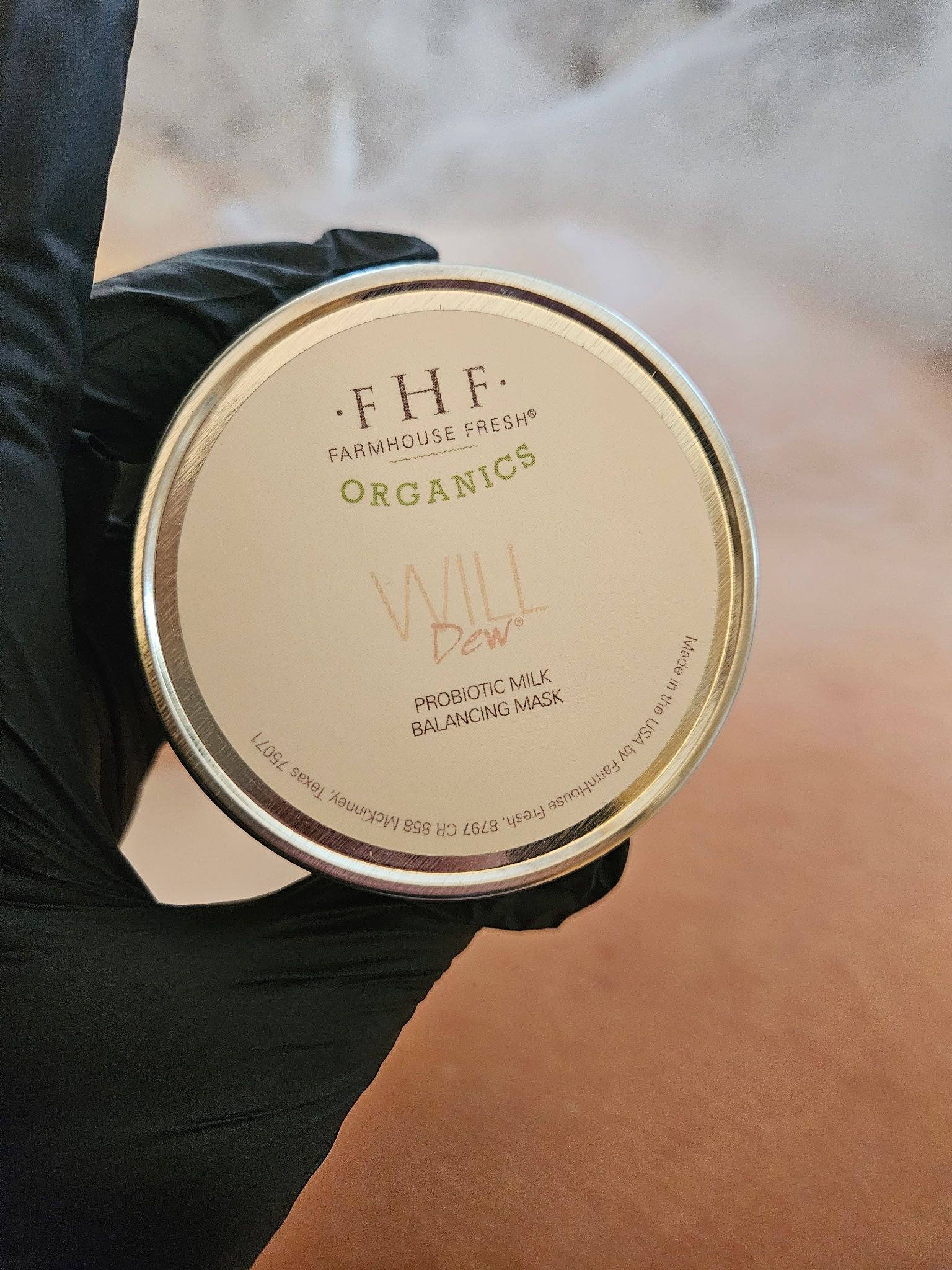 A black-gloved hand holding a round, gold-lidded product container; label reads 