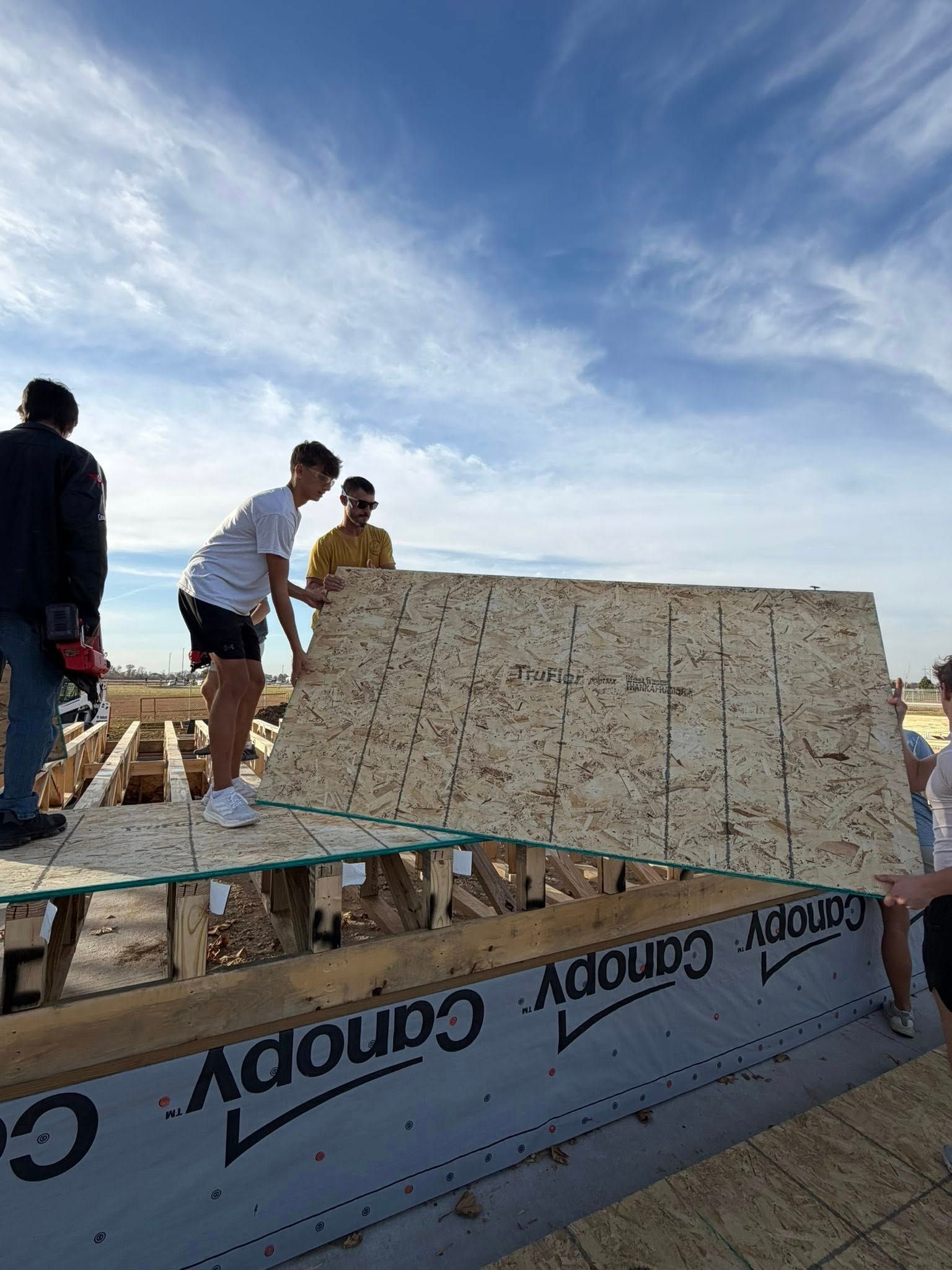 People working together to install a large panel of plywood onto the floor joists of a new building frame under a blue sky.