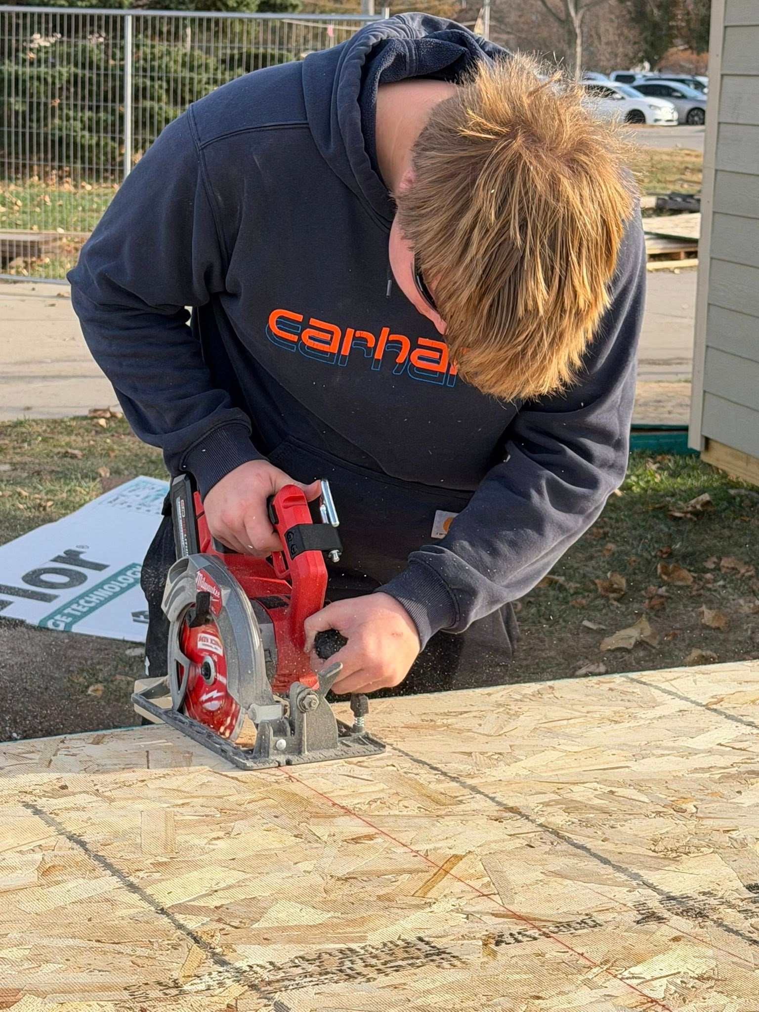 A person wearing a Carhartt hoodie uses a red circular saw to cut a piece of plywood on a construction site.