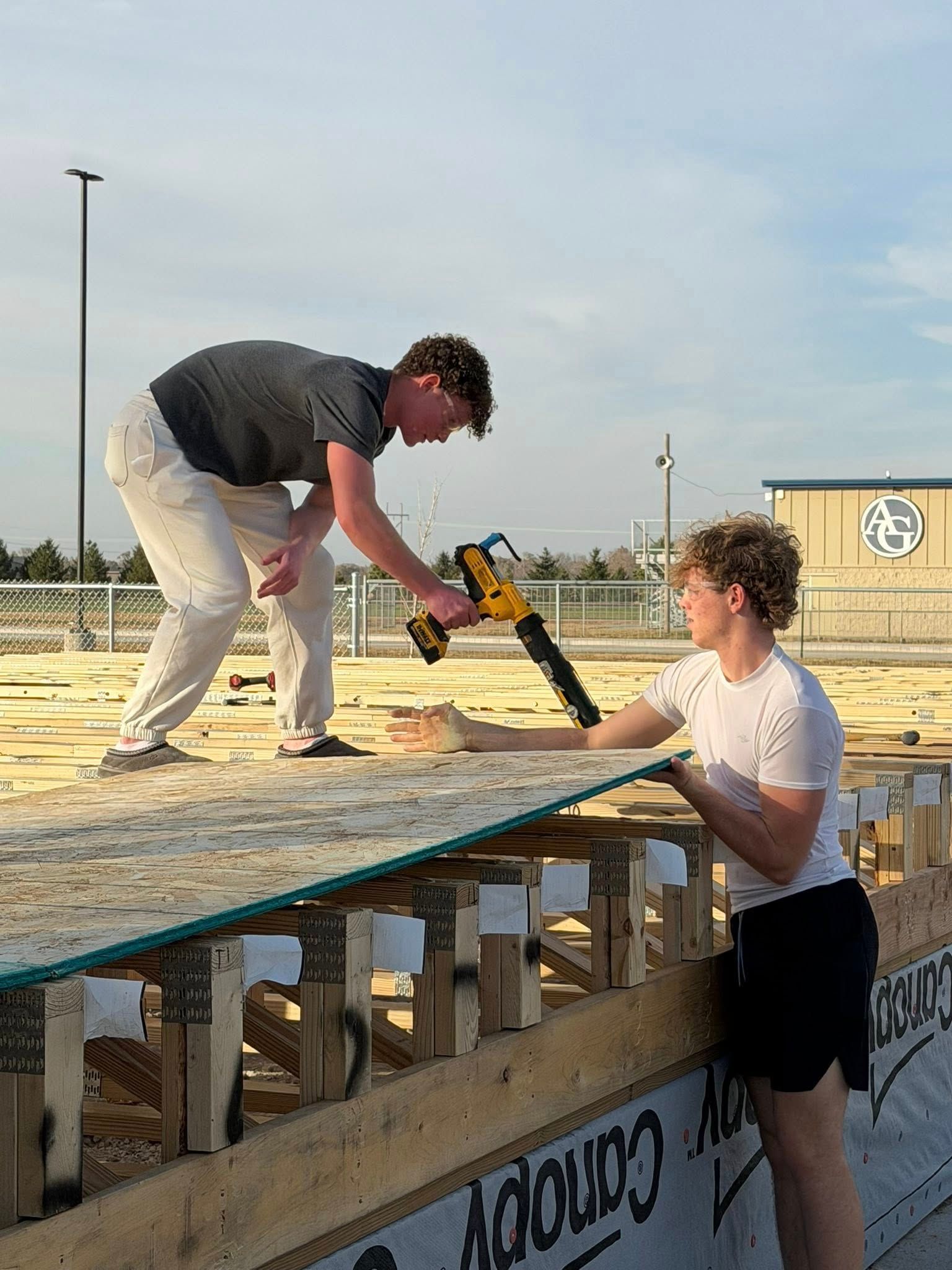 Two people work on a wooden construction frame on a bright day; one uses a yellow power tool to fasten a plywood sheet.