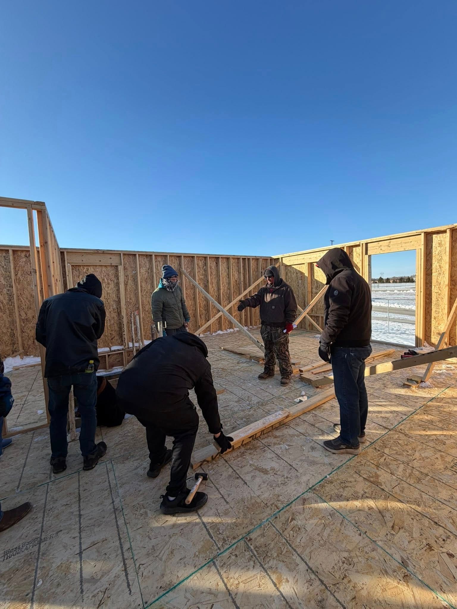 Five people in winter gear working on the wooden frame of a building under a clear blue sky.