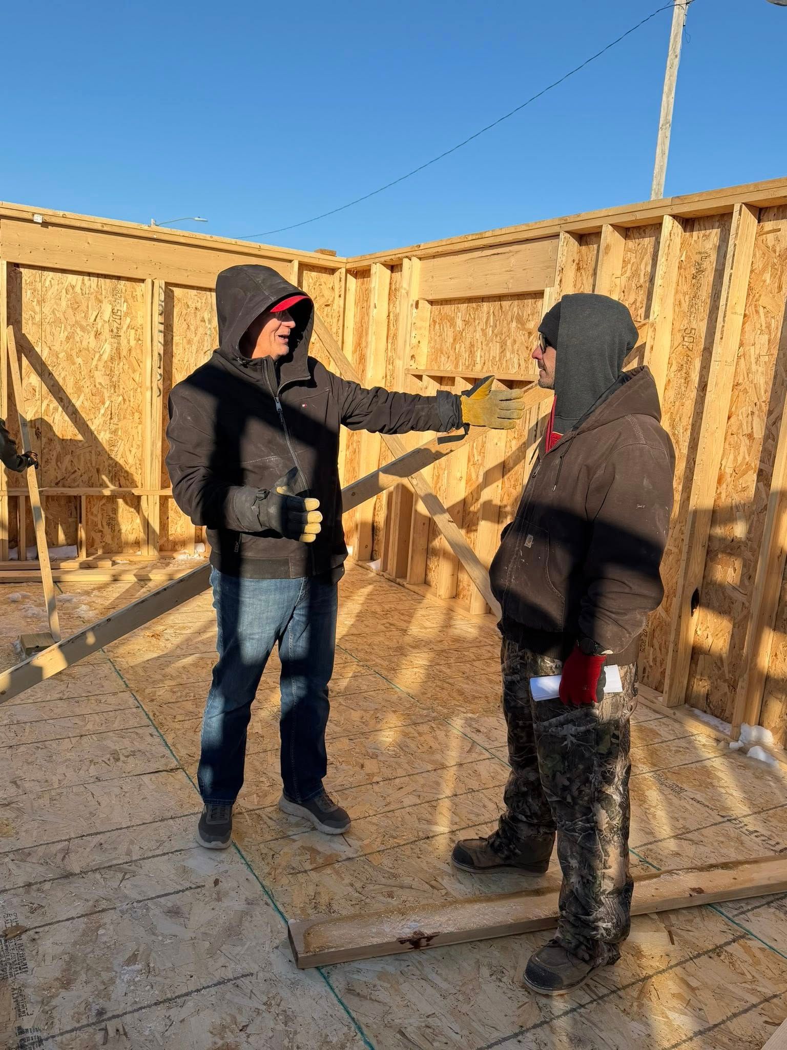 Two people wearing hooded jackets stand inside a wooden wall frame under construction, talking in bright sunlight.