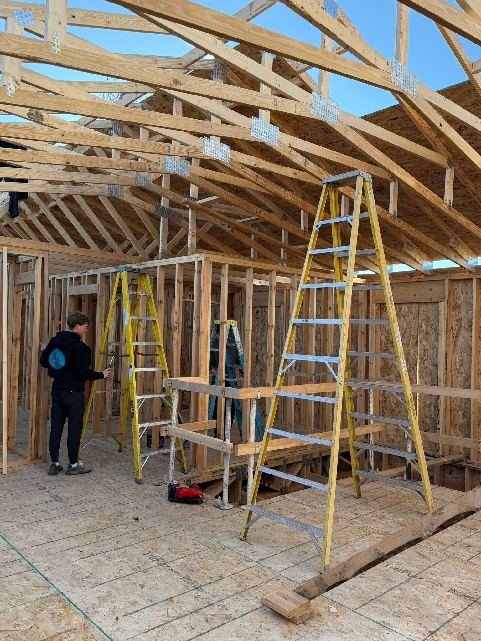 A person stands inside the wood-framed interior of a house under construction, with two yellow ladders nearby.