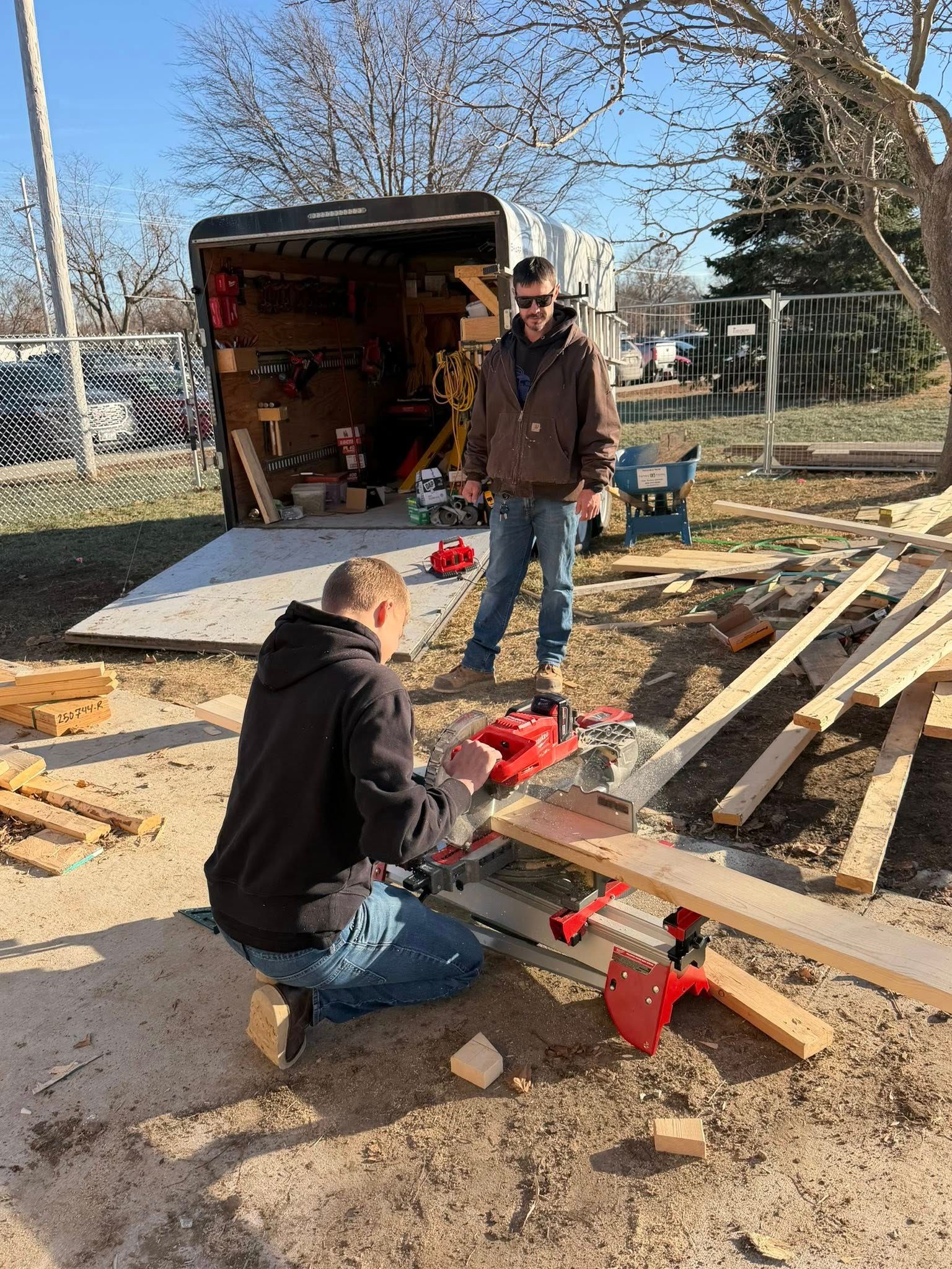 Two people work with wood and tools on a construction site outside a trailer on a sunny day.