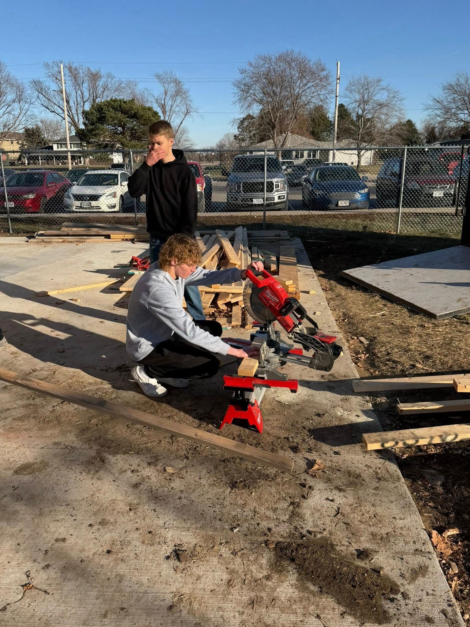 Two people work outside on a construction project, one operating a red miter saw on a concrete surface.
