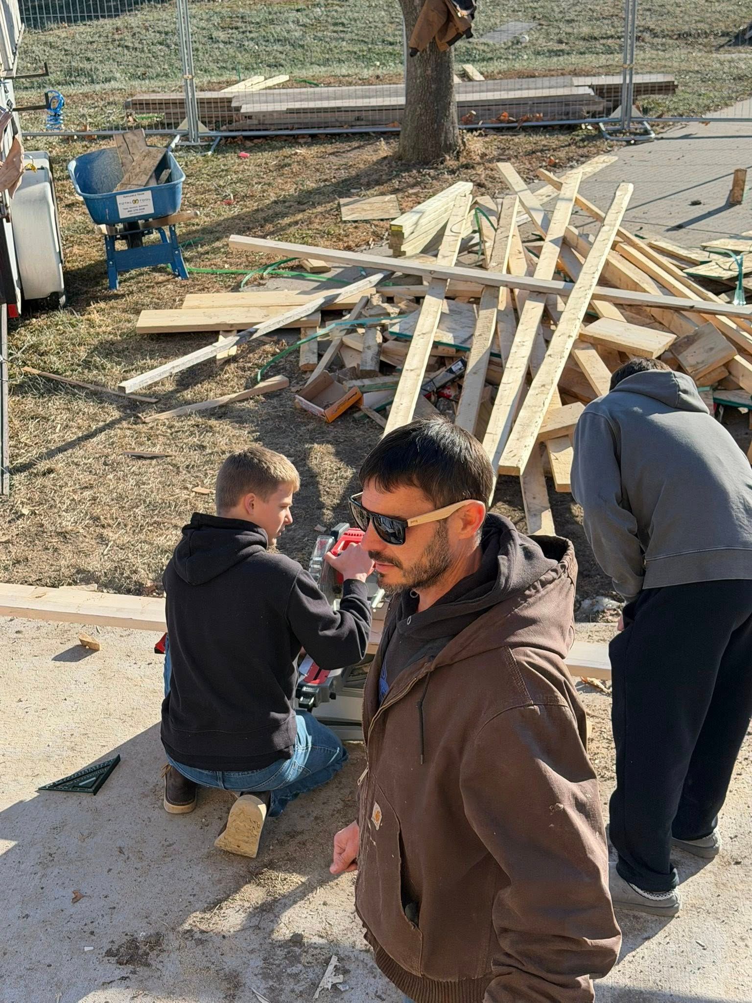 Three people work at a construction site with piles of lumber, a blue wheelbarrow, and scaffolding in the background.