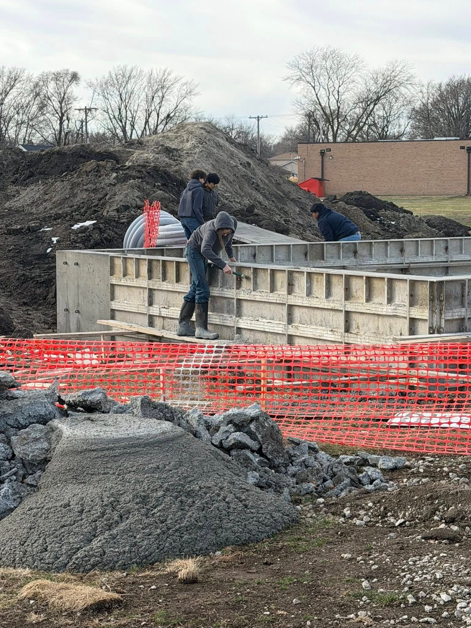 Construction workers stand on a concrete foundation at a building site, with piles of dirt and orange fencing nearby.