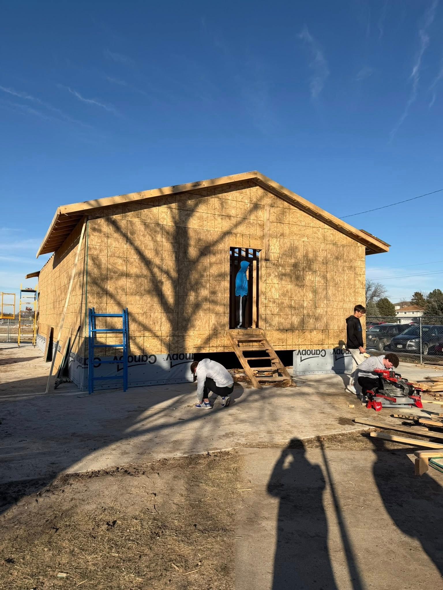 People work on the exterior of a wood-framed house under a clear blue sky.