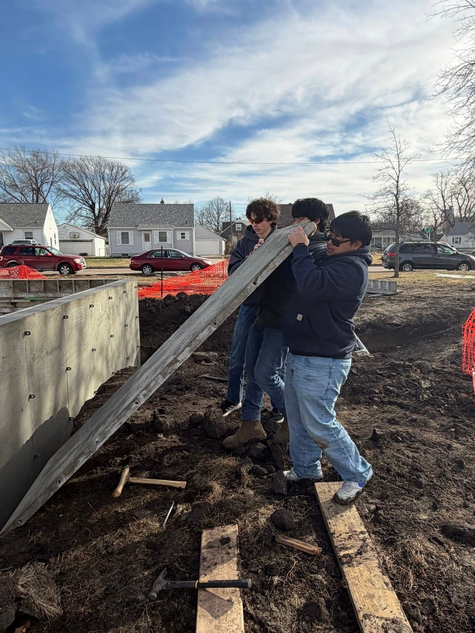 Three people work together to lift a long, heavy wooden beam against a concrete wall at an outdoor construction site.