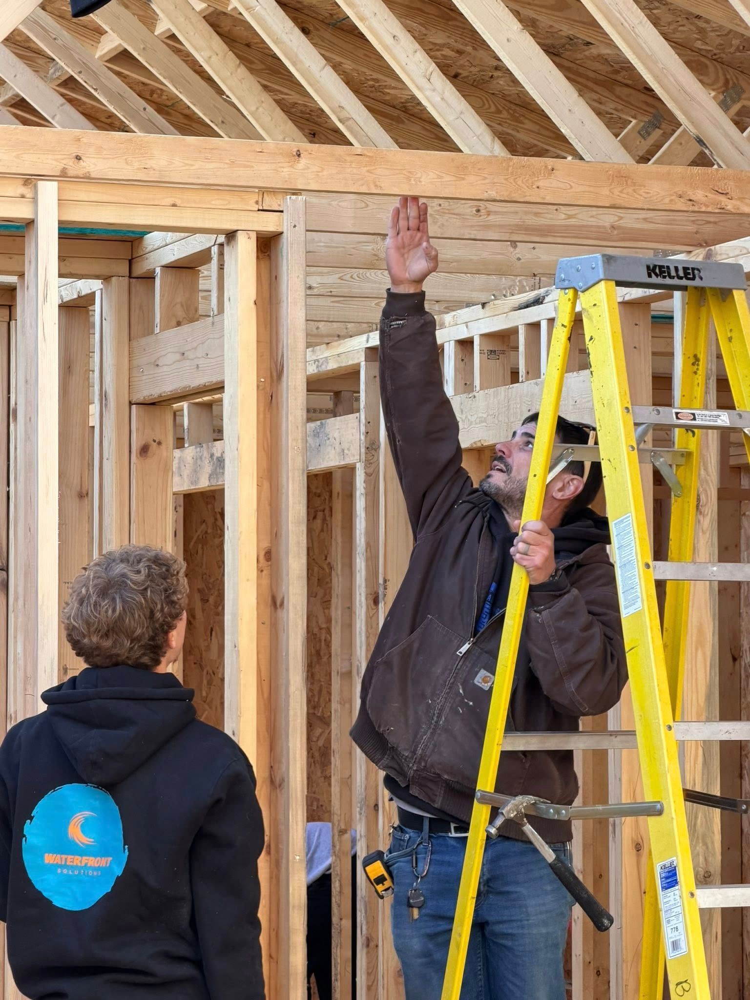 A worker stands on a yellow ladder, pointing at the wooden framing of a building under construction, observed by another.