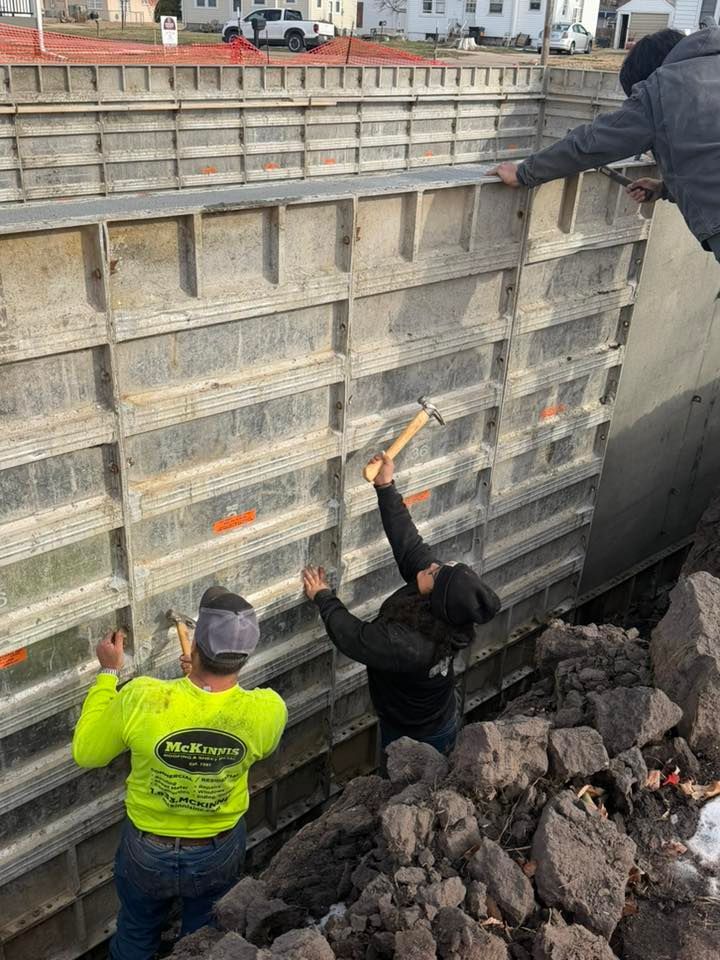 Construction workers in a trench assemble metal concrete forms for a foundation wall using hammers.