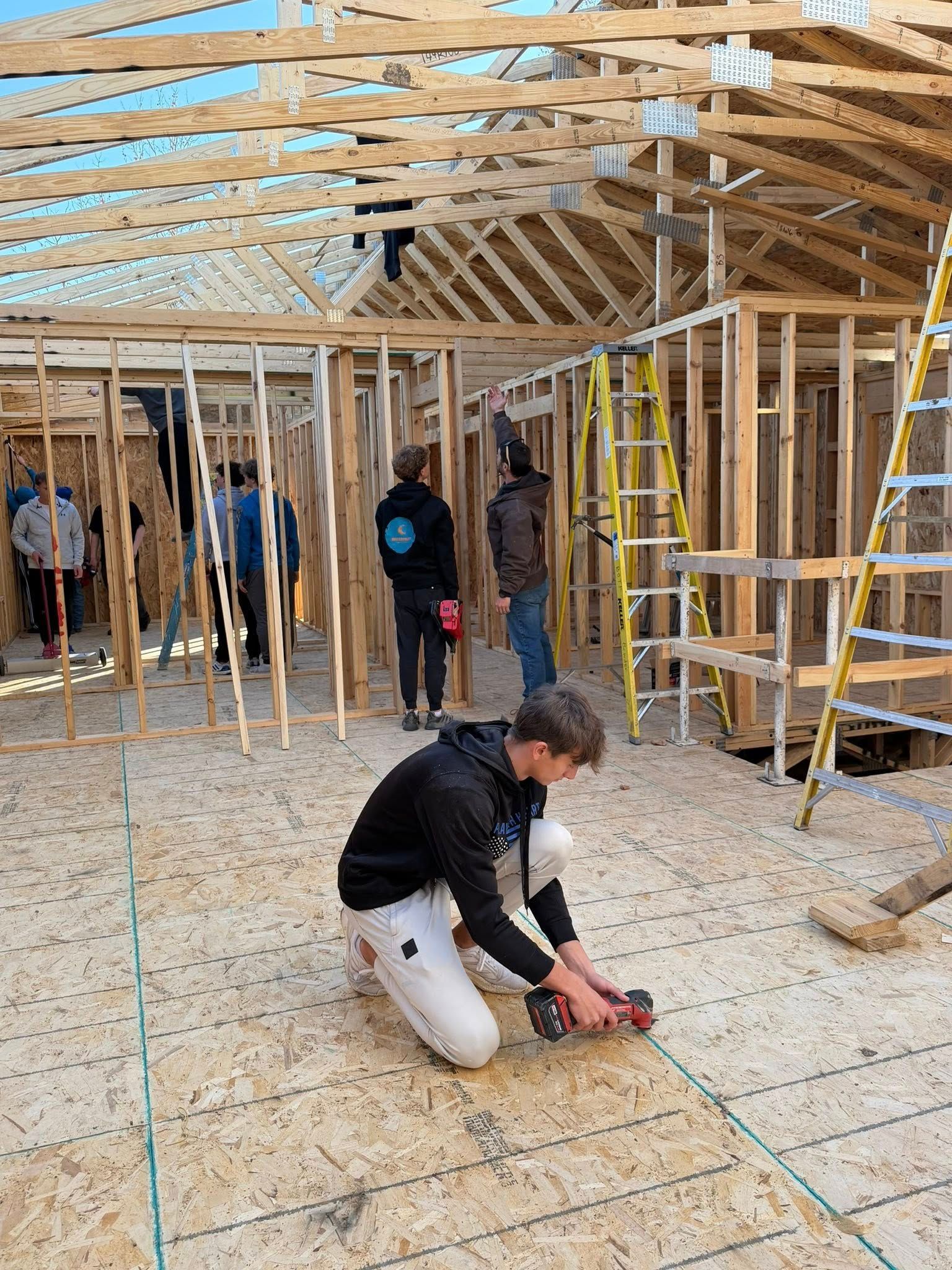 Construction workers building the wooden frame of a house interior, with one individual kneeling to use a power drill.