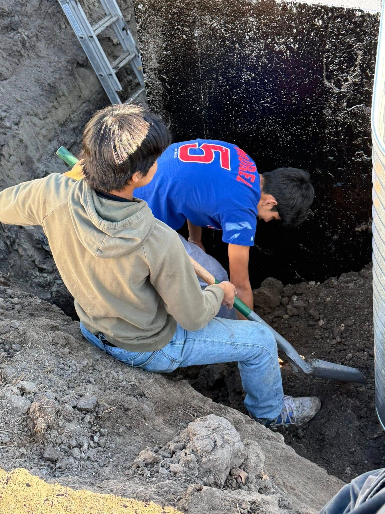 Two people stand in a deep trench, using shovels to dig into the dirt near a ladder.