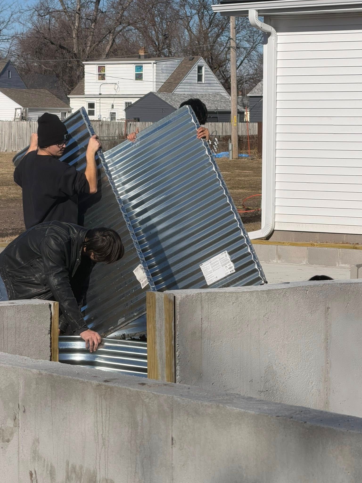 Two people in black clothing maneuver a large, corrugated metal panel into position against a concrete foundation.