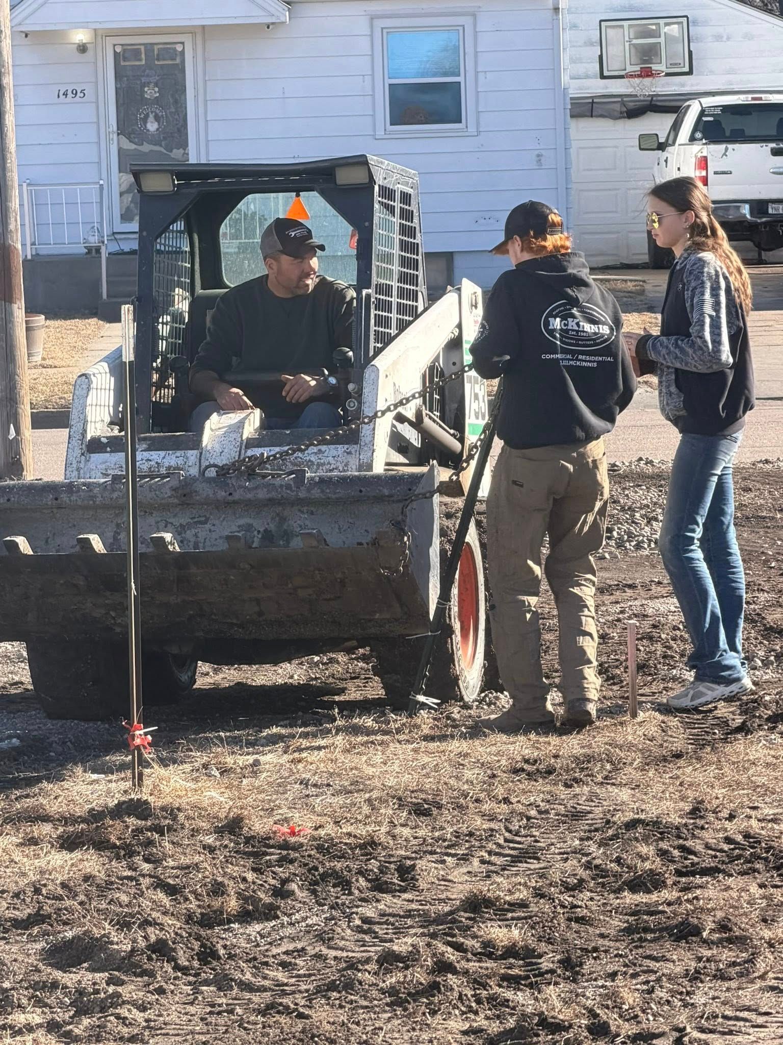 A person sits in a skid-steer loader while two others stand nearby in a muddy lot in front of a house.