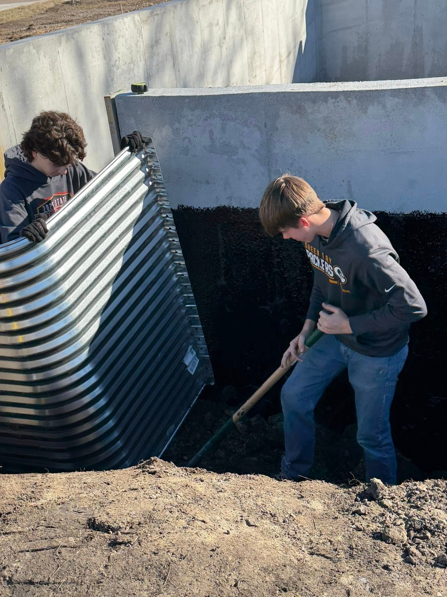 Two people working in a trench, one positioning a corrugated metal window well while the other digs with a shovel.