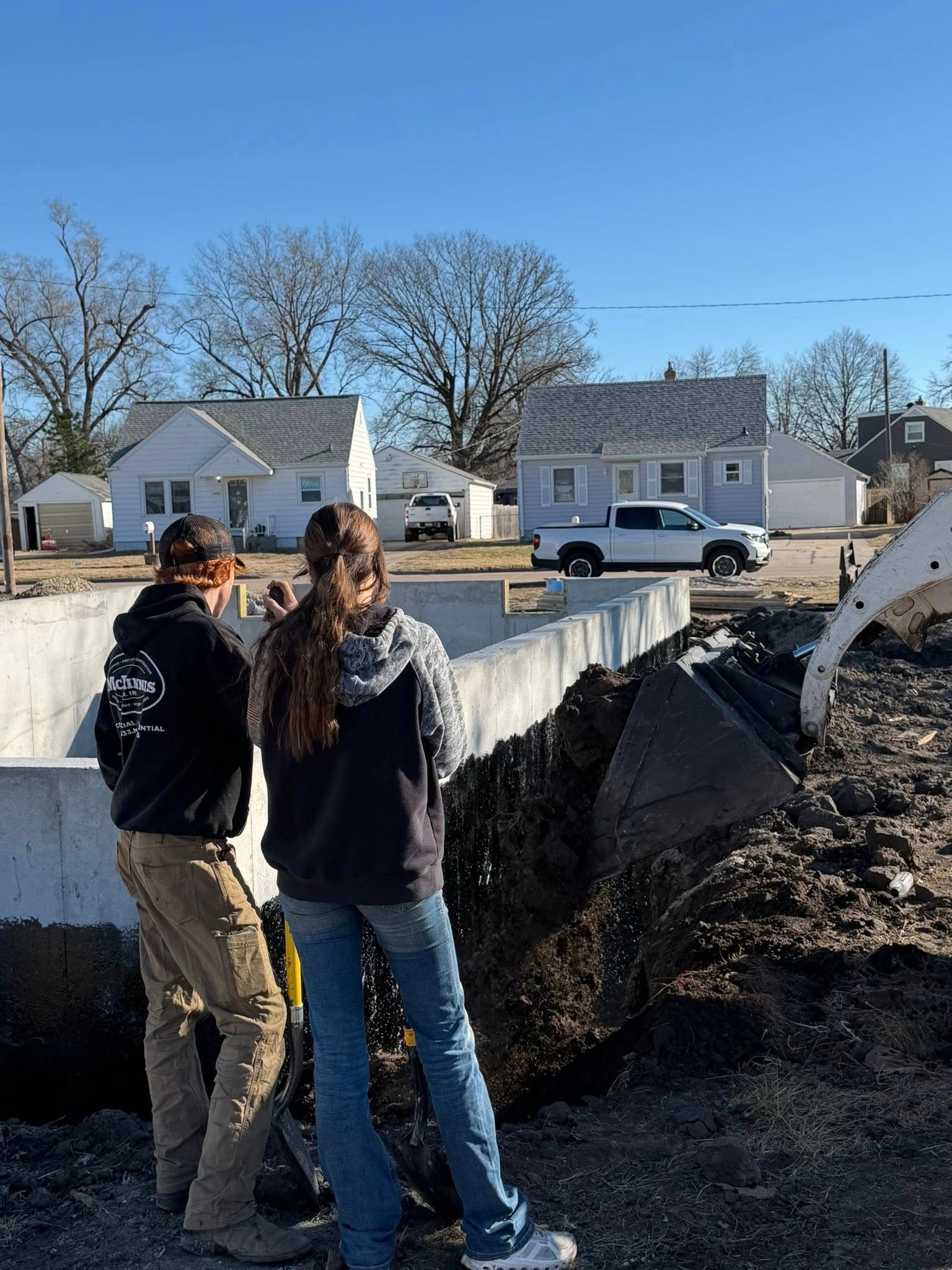 Two people in hoodies and jeans stand on a construction site, observing an excavator digging near concrete foundations.