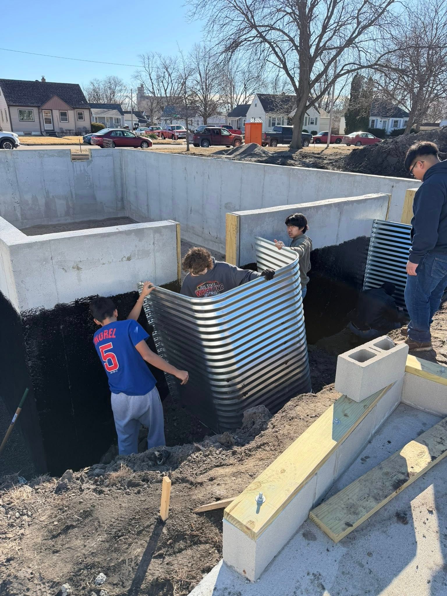 Three people install a corrugated metal window well against a concrete foundation at a construction site.