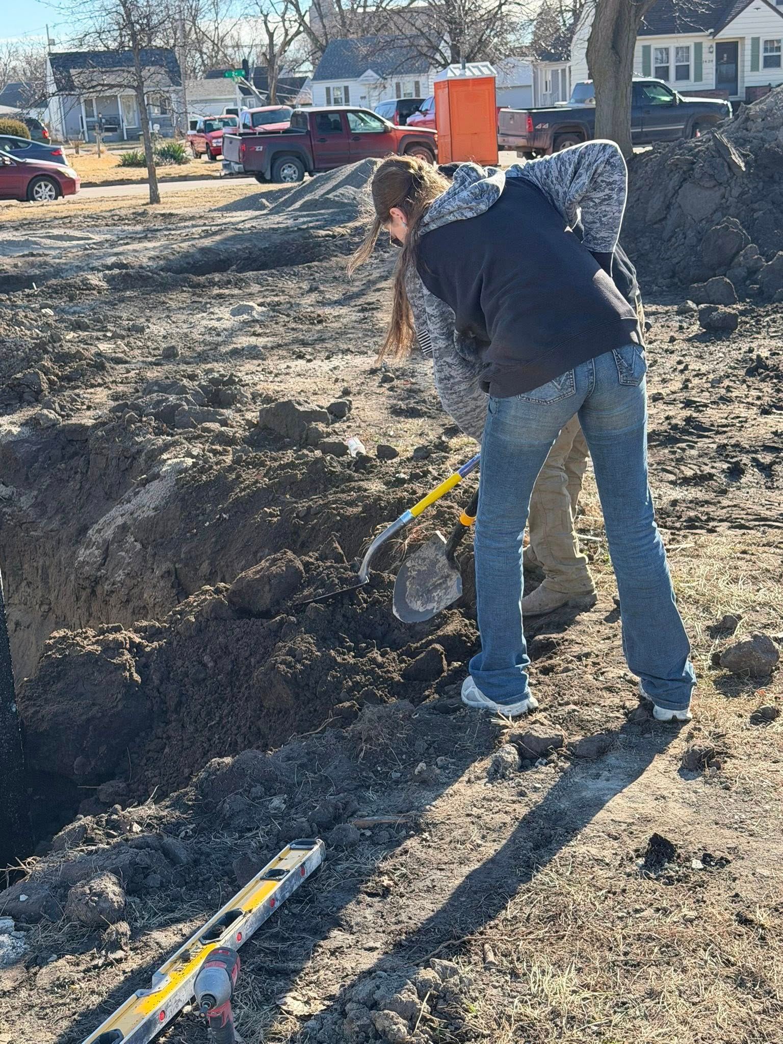 A person in a dark jacket and blue jeans uses a shovel to dig in a dirt construction site with a level nearby.
