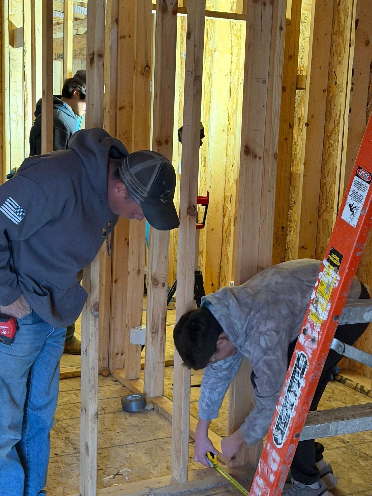 Two construction workers measure a wall frame inside an unfinished wood-framed building.