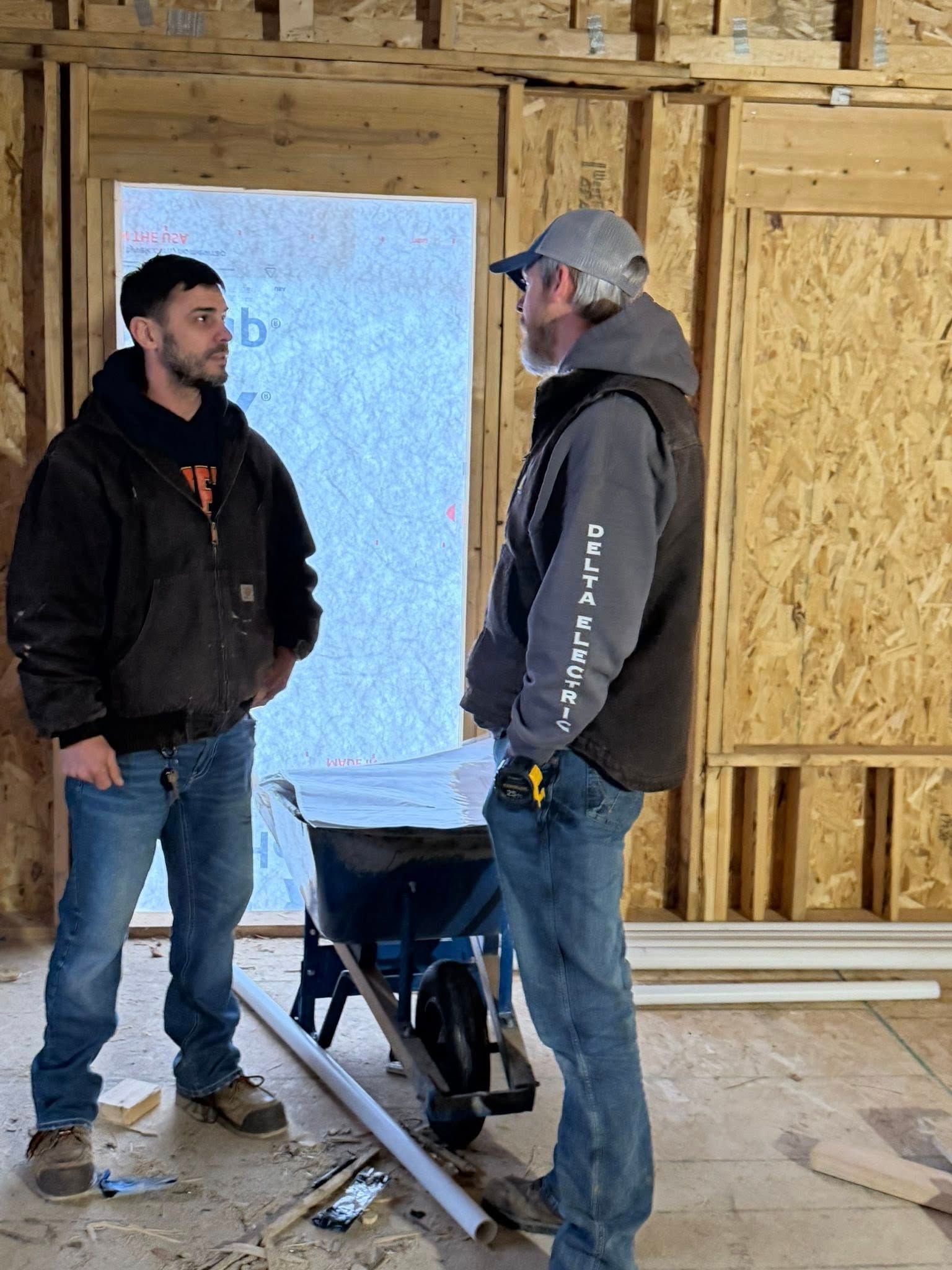 Two people wearing work clothes and hoodies standing and talking inside an unfinished building frame with a wheelbarrow.